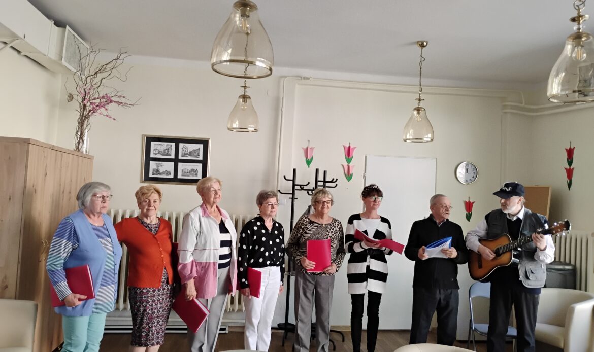 Zespół składający się z sześciu pań i dwóch panów stoi ze śpiewnikami w dłoniach. Mężczyzna po prawej stronie gra na gitarze.