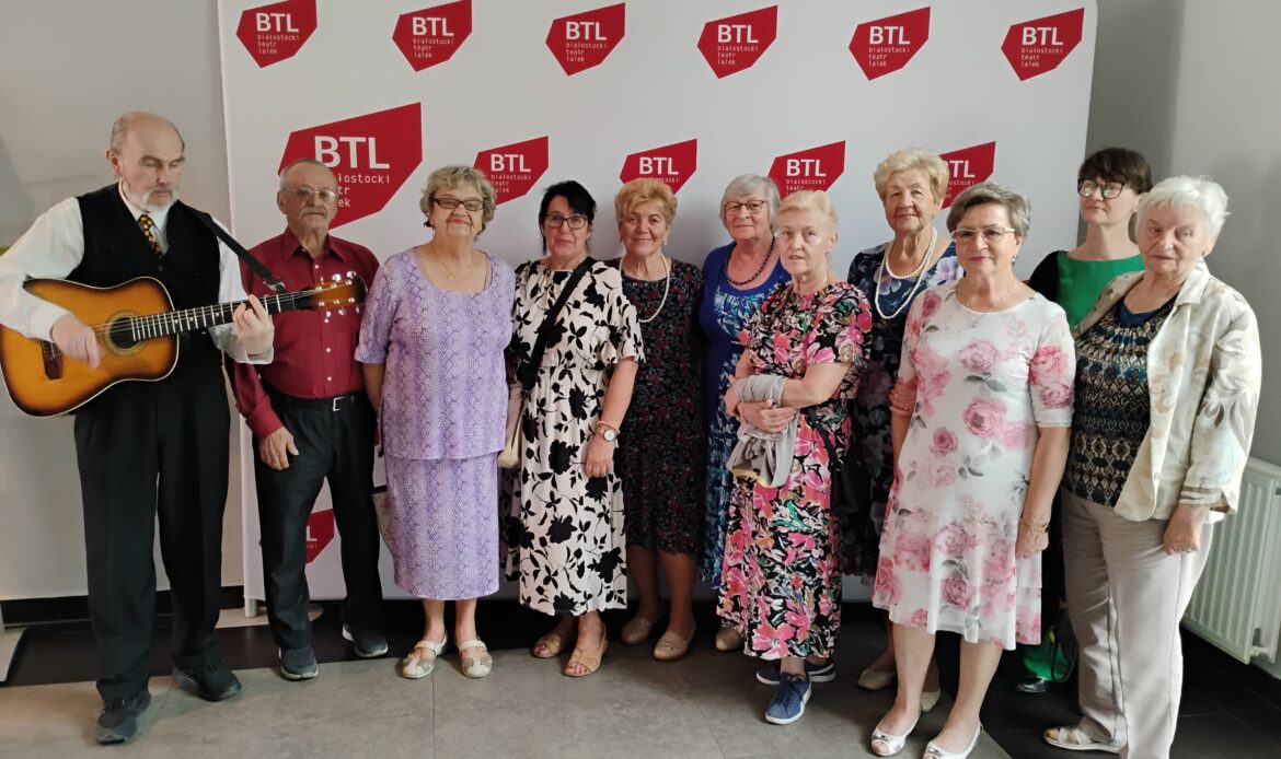 .Jedenastoosobowa grupa seniorów na tle ścianki ze skrótem Białostocki Teatr Lalek.