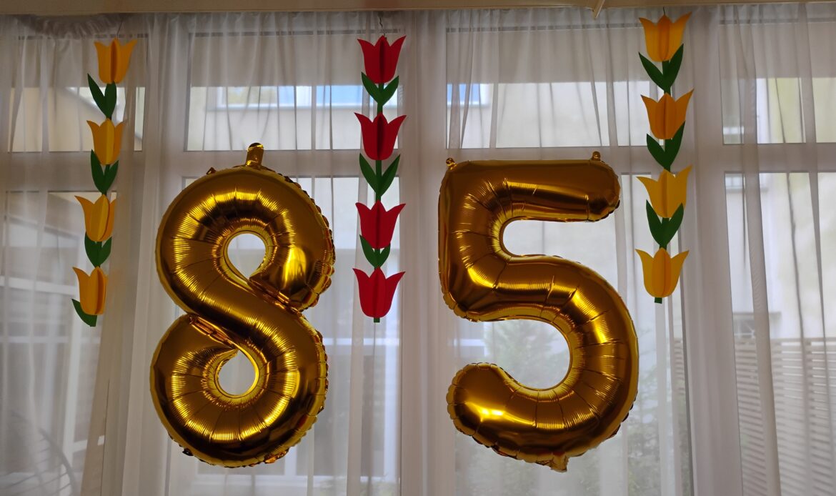 Dwa balony w kształcie liczby 85 zawieszone na firance.