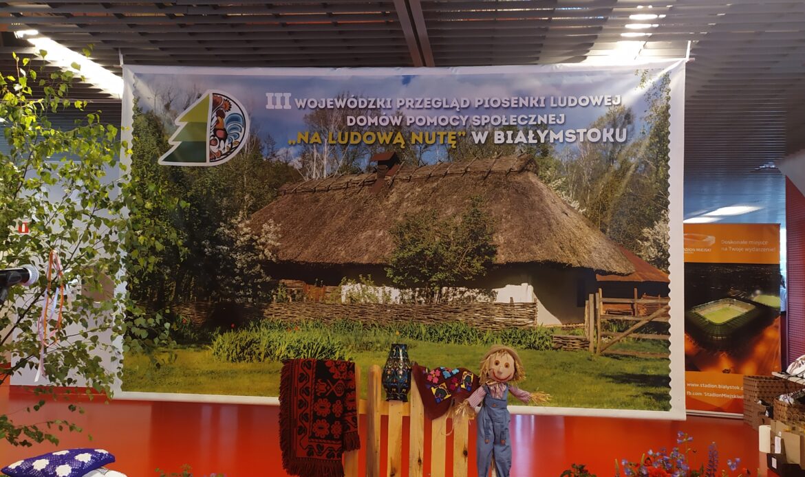 Baner przedstawiający wiejską chatę i informacje dotyczące przeglądu. Na pierwszym planie ustawiony płot, na nim wiejska kapa, dzban, chusta i obok strach na wróble.
