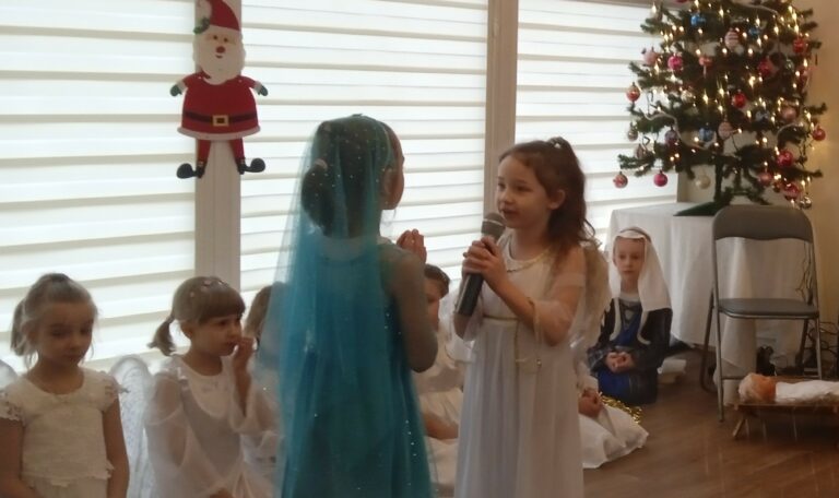 Dwie dziewczynki stoją na środku sali zwrócone do siebie, jedna jest w niebieskim, a druga w białym stroju. Dziewczynka przedstawiająca anioła mówi do mikrofonu.