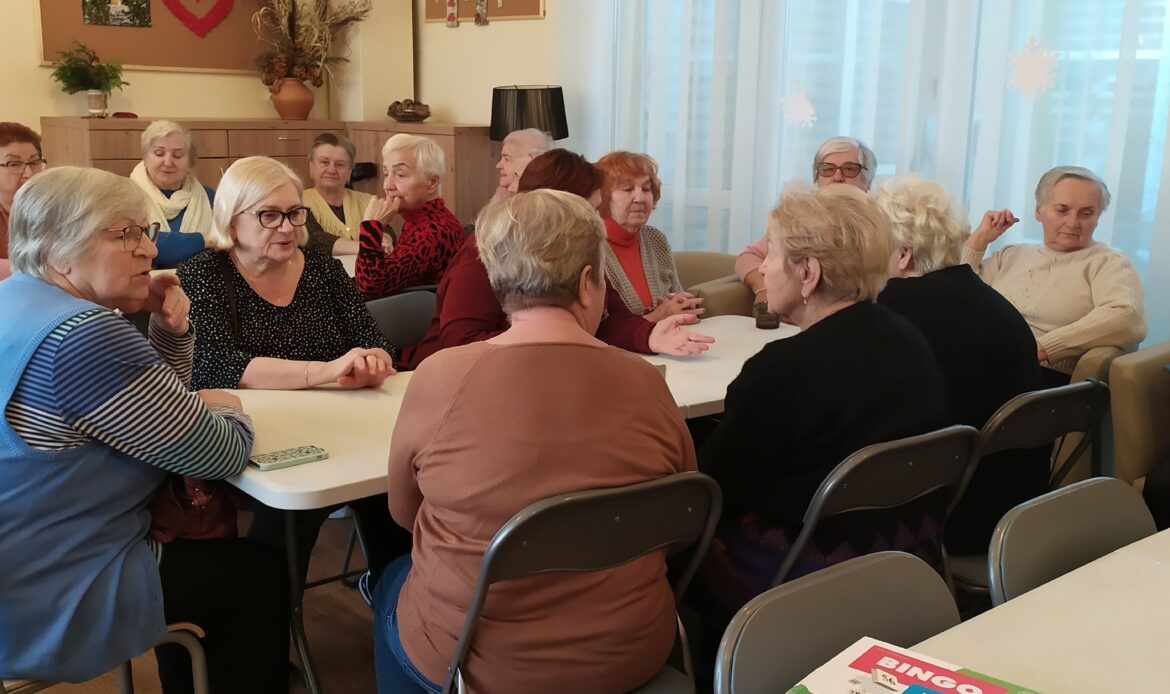 Seniorzy siedzą przy stołach ustawionych w trzech rzędach. W prawym dolnym rogu na blacie leży opakowanie z grą Bingo.