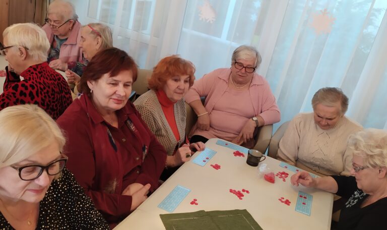 Grupa seniorów skupiona wokół stołu, na którym leżą małe, błękitne plansze do gry i czerwone żetony.