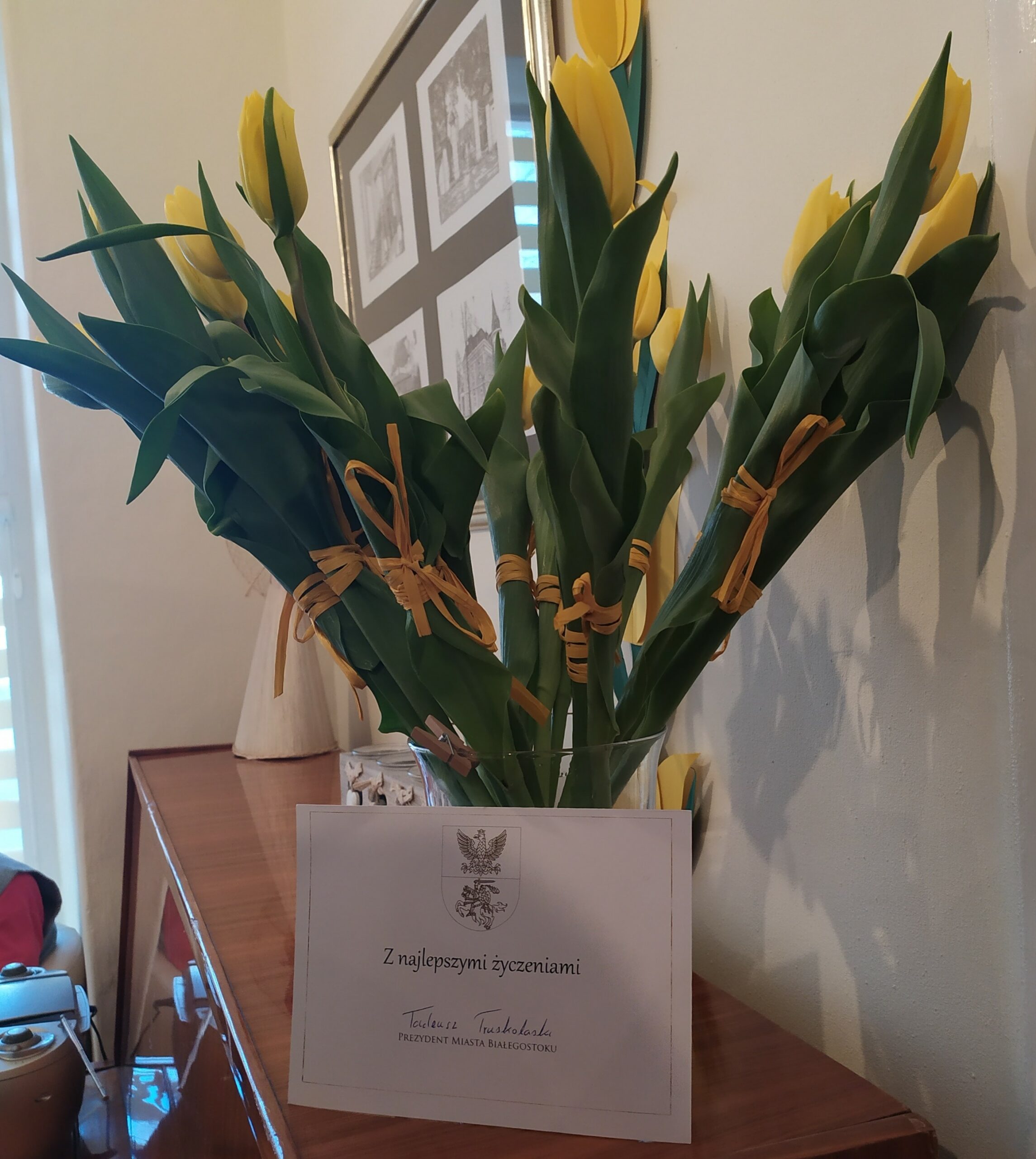 Bukiet żółtych tulipanów w szklanym wazonie i kartka z napisem: "Z najlepszymi życzeniami Tadeusz Truskolaski Prezydent Miasta Białegostoku"
