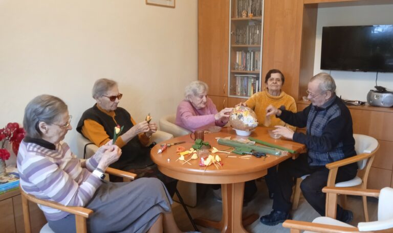 Grupa pięciu osób siedzi przy stole. Trzy oklejają balon brązowym papierem, a dwie robią krokusy z krepiny.