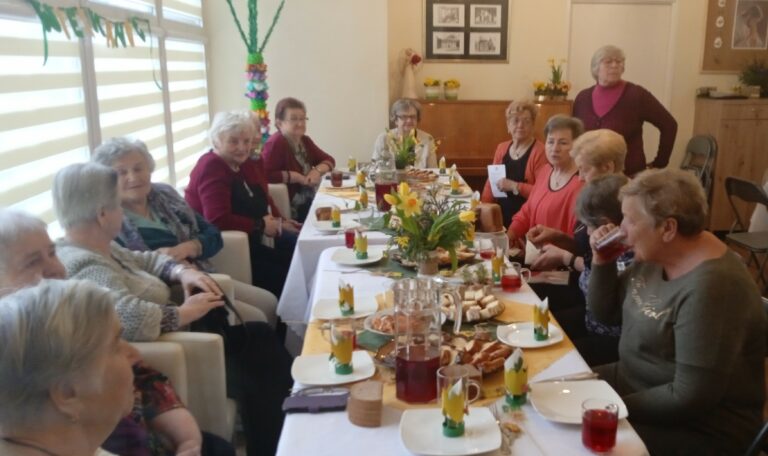 Grupa seniorów ze świetlicy z ul. Nowogródzkiej siedzi po obu stronach świątecznie udekorowanego i zastawionego potrawami stołu.