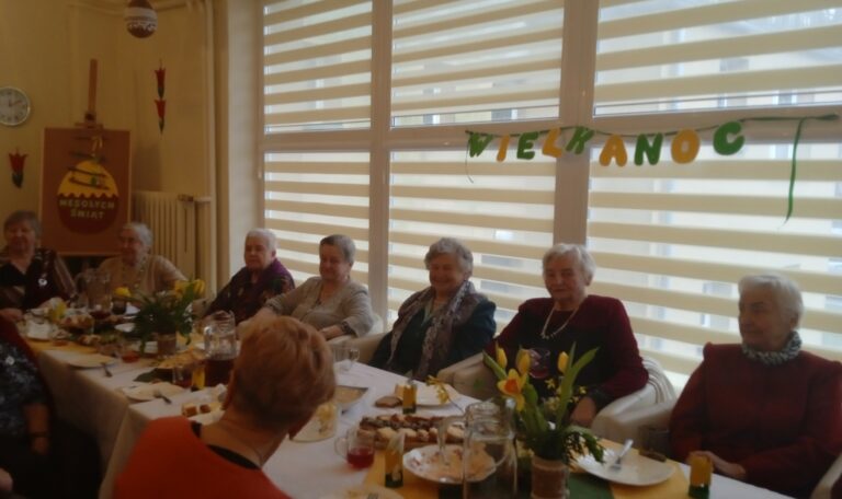 Siedem pań siedzi przodem przy stole, w tle okno i dekoracje z napisami Wielkanoc i Wesołych Świąt.